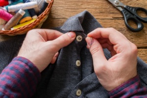 A person sewing a button onto a gray shirt with thread, needles, and sewing