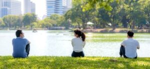 Three people sitting on the lakeside in a city park, facing the water