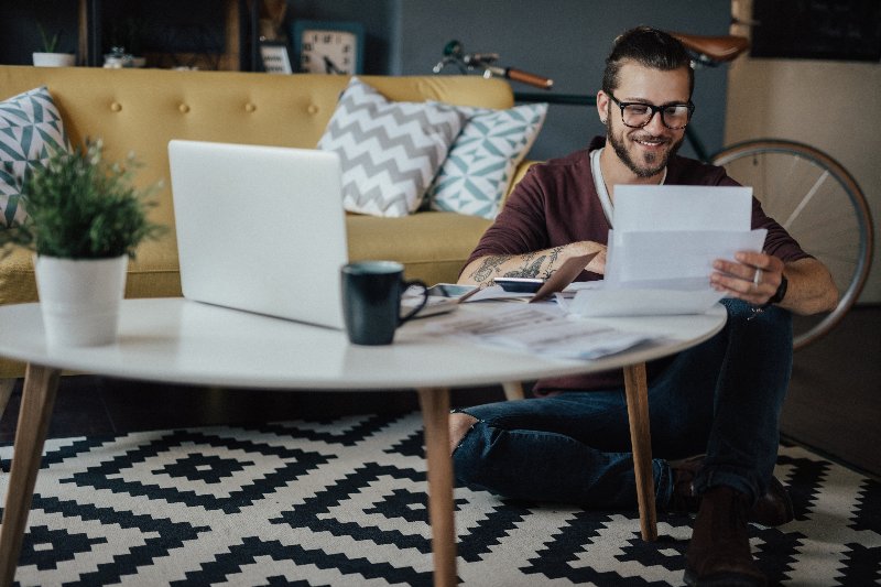 A millennial man sitting on the floor, smiling while looking at papers by Century Support Services