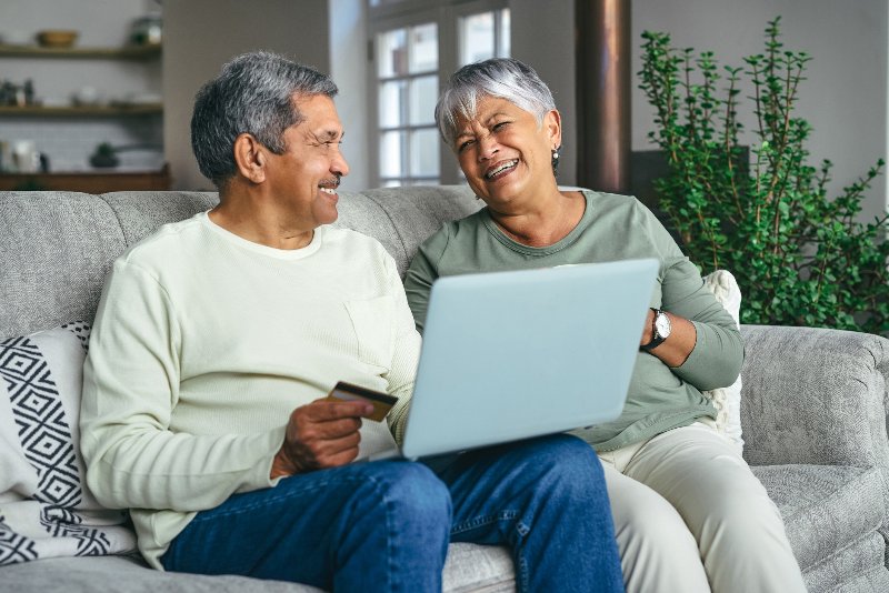 An elderly couple sitting on a couch with a laptop and smiling by Century Support Services
