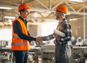 Two construction workers shaking hands in an industrial setting with machinery behind them