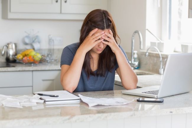 Person sitting at a kitchen counter with a laptop and bills, appearing stressed