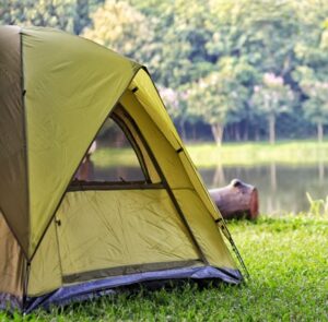 Camping tent on grass beside a calm lake with trees in the background.
