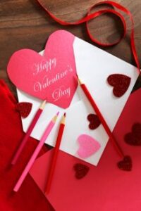 Valentine’s card with pink heart, colored pencils, and red glitter hearts on a table