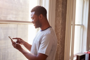 Person standing near a window using a smartphone with bookshelves
