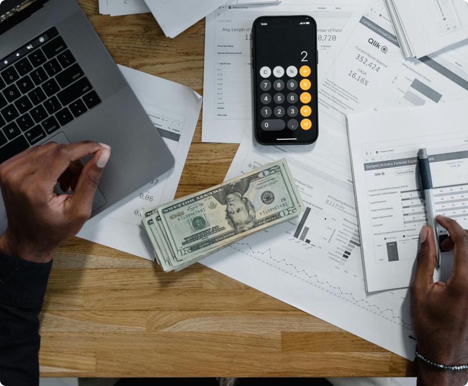 Hands reviewing financial documents with a calculator app, cash, and a laptop on a table